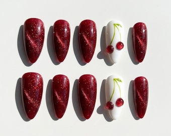 May include: A set of ten almond-shaped press-on nails. Eight nails feature a deep red cat-eye magnetic polish with a shimmering, reflective finish. Two nails are white with a hand-painted green stem and two small, raised red cherry accents.