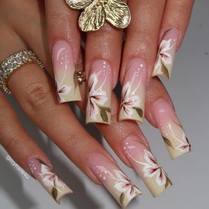 May include: Long, square-shaped acrylic nails with a floral design. The nails feature a gradient from a light pink at the cuticle to a beige tip, with white and maroon flower accents and green leaves. A gold flower ring and a diamond ring are also visible.