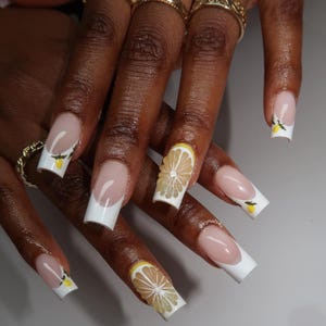 May include: A set of long, square-shaped acrylic nails with a French tip design. The nails feature a white tip, a nude base, and lemon slice and lemon sprig nail art. Gold rings adorn the fingers.
