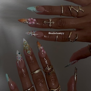 May include: Close-up of long, pointed acrylic nails with various designs. Some nails feature floral art, others have gems, and some have a French tip design. Gold rings adorn the fingers. The text "@nailedxamyy" is visible.