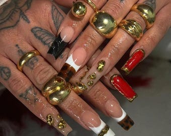 May include: Close-up of hands adorned with gold rings of various designs. The nails feature a range of styles, including French tips with black, white, and leopard print accents, and red nails with gold trim. Some nails are embellished with gold charms.