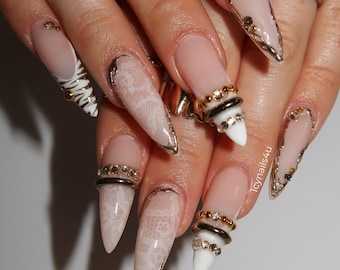 May include: Close-up of long, pointed acrylic nails with various designs. Some nails feature a nude base with gold accents, lace patterns, or zebra stripes. Several gold rings adorn the fingers, adding to the glamorous aesthetic. The text "1Cynails4u" is visible.