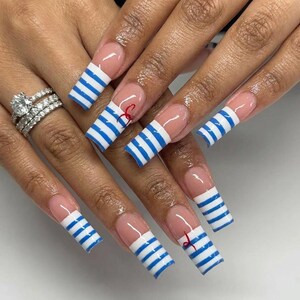 Op de afbeelding: Lange, vierkante kunstnagels in een French manicure-stijl met blauwe en witte horizontale strepen op de uiteinden. Twee van de nagels hebben een klein, delicaat rood accent in cursief schrift. Er wordt een zilverkleurige ring met heldere stenen gedragen.