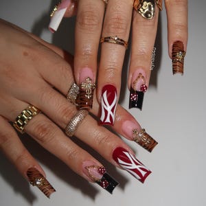 Puede incluir: Primer plano de uñas manicuradas con diversos diseños. Algunas uñas están pintadas de rojo con detalles blancos, otras presentan rayas de tigre y algunas son negras con lunares rojos. Anillos y joyas de oro adornan los dedos.