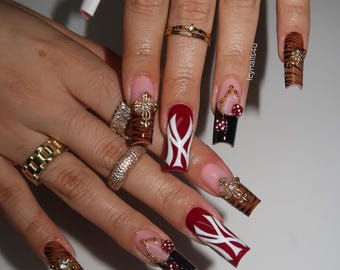 May include: A close-up of manicured nails with various designs. Some nails are painted red with white accents, others feature tiger stripes, and some are black with red polka dots. Gold rings and jewelry adorn the fingers.