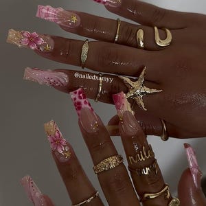 Puede incluir: Primer plano de una mano con uñas largas y decoradas. Las uñas presentan diseños rosas y blancos, incluyendo acentos florales, patrones y adornos dorados. Varios anillos dorados adornan los dedos, incluyendo un anillo de estrella de mar.