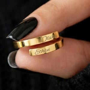 May include: Two gold rings, each engraved with a name, are wrapped around a finger. The top ring reads "Olivia", the bottom ring reads "Ethan". The rings are shown against a black background.