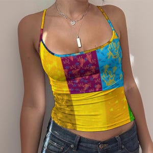 Könnte beinhalten: Ein ärmelloses Tanktop mit einem lebendigen Patchwork-Design aus leuchtend gelben, tiefvioletten und himmelblauen Feldern. Der Stoff zeigt ein wiederkehrendes Muster aus kleinen Sternen und blattartigen Formen, abgerundet durch dünne Träger und einen Rundhalsausschnitt.