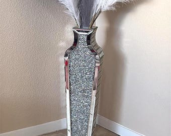 Puede incluir: Un jarrón decorativo alto con exterior espejado y una sección central brillante. Contiene una mezcla de plumas blancas y grises. El jarrón está colocado en una esquina contra una pared neutra. El jarrón mide aproximadamente 100 cm de alto.