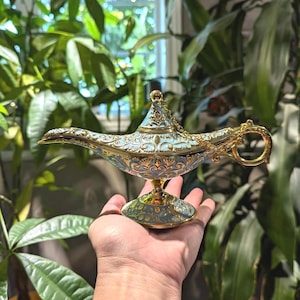 Puede incluir: Una lámpara de genio decorativa con un asa y un pico dorados, y un cuerpo dorado y azul. La lámpara tiene detalles intrincados y una pequeña tapa redonda. La lámpara se sostiene en una mano.