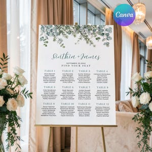 May include: A white wedding seating chart sign on a gold easel featuring green eucalyptus leaf illustrations at the top. The text reads Sinthia + James, September 25, 20XX, Find Your Seat, and lists guest names organised by tables one through twelve.
