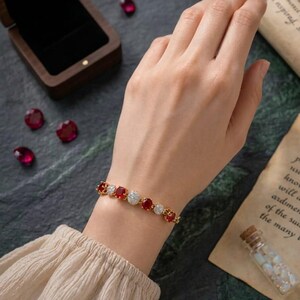 Op de afbeelding: Een gouden armband met afwisselend ronde rode edelstenen en heldere witte stenen. De armband wordt om de pols gedragen, met een houten juwelendoos, losse rode stenen en een klein glazen flesje met witte kralen in de nabijheid.