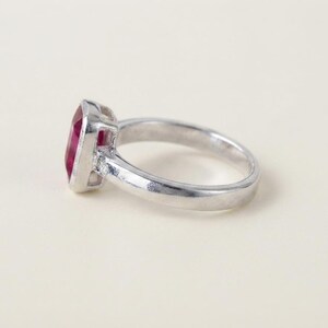 May include: A silver-toned ring featuring a rectangular, deep pink gemstone set in a bezel setting. The band is smooth and polished, with a small, clear accent stone visible on the side where the band meets the central gemstone setting.