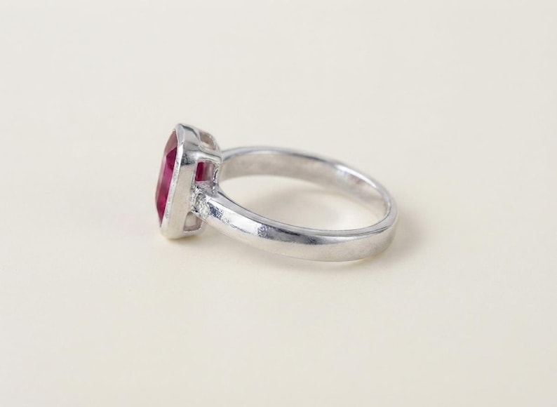 May include: A silver-toned ring featuring a rectangular, deep pink gemstone set in a bezel setting. The band is smooth and polished, with a small, clear accent stone visible on the side where the band meets the central gemstone setting.
