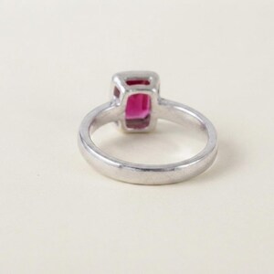 May include: A silver-toned ring featuring a rectangular, vibrant pink gemstone set in a smooth metal bezel. The band is plain and polished, showcasing a clean, simple design that highlights the central stone from a side-angle perspective.