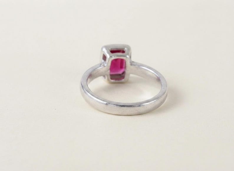 May include: A silver-toned ring featuring a rectangular, vibrant pink gemstone set in a smooth metal bezel. The band is plain and polished, showcasing a clean, simple design that highlights the central stone from a side-angle perspective.