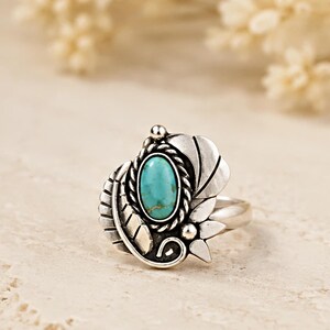 May include: A silver ring featuring an oval turquoise stone set in a twisted rope border. The setting is decorated with metal leaf-like shapes and small spheres, creating a detailed design that surrounds the central blue-green gemstone.