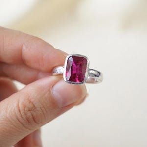 May include: A silver ring featuring a rectangular, faceted, deep pink gemstone set in a smooth bezel. The metal band has a textured, hammered finish along the sides, providing a contrast to the polished setting of the vibrant, dark pink stone.