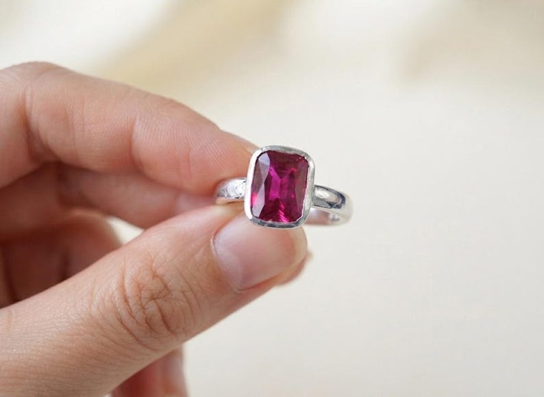 May include: A silver ring featuring a rectangular, faceted, deep pink gemstone set in a smooth bezel. The metal band has a textured, hammered finish along the sides, providing a contrast to the polished setting of the vibrant, dark pink stone.