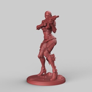 May include: A red-brown figurine of a female character in a combat pose, holding a rifle. The figure is wearing armor and high-heeled boots, standing on a circular base. The overall design suggests a science fiction or fantasy theme.