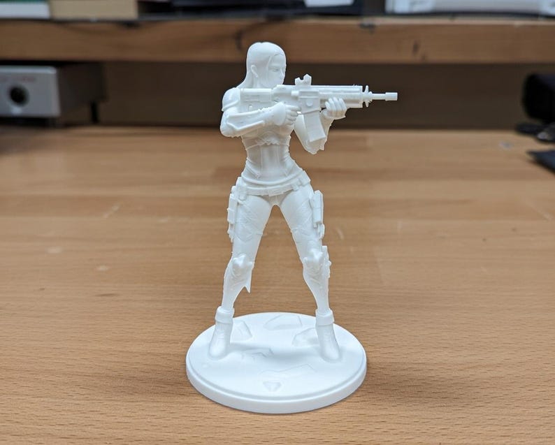 May include: A white 3D-printed figurine of a female character holding a rifle. The figure is standing on a circular base. The character is wearing a form-fitting outfit with armor details.