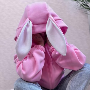 Op de afbeelding: Een roze hoodie met grote konijnenoren en een witte binnenkant. De hoodie heeft een ritssluiting en wordt gedragen met lichtblauwe jeans. De mouwen zijn bij de polsen gerimpeld.