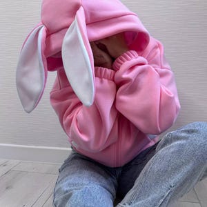 Op de afbeelding: Een roze konijnenhoodie met lange, slappe oren en een witte binnenkant. De hoodie heeft een ritssluiting en geribde manchetten. De persoon draagt een blauwe spijkerbroek. De outfit is casual en speels.