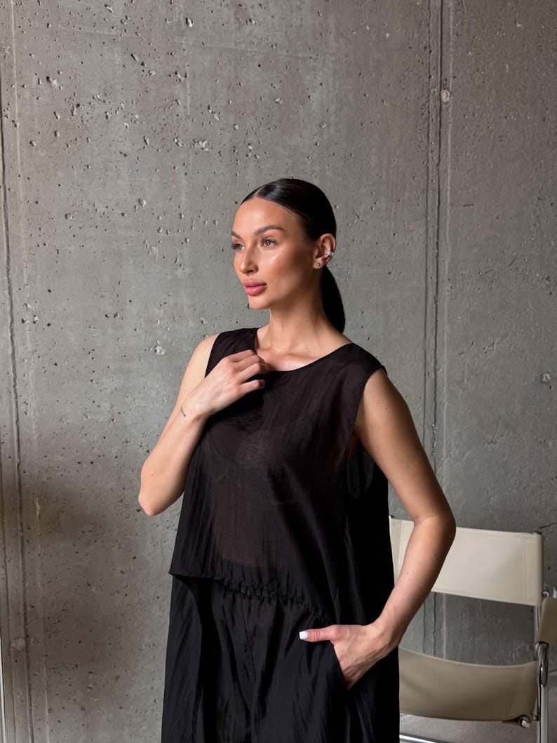May include: A sleeveless black top with a round neckline and a slightly sheer, textured fabric. The garment features a loose, relaxed fit with a straight hemline that falls at the hip, showcasing a subtle sheen and a lightweight, breathable construction.