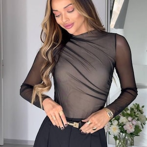 Könnte beinhalten: Schwarzes, durchsichtiges Langarm-Top mit hohem Ausschnitt und Falten. Kombiniert mit schwarzer Faltenhose und einem goldenen Gürtel. Das Outfit ist mit goldenen Ringen und einer Uhr ausgestattet.