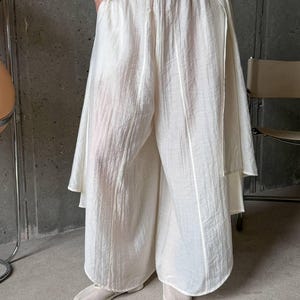 May include: Wide-leg trousers in an off-white color featuring a textured, crinkled fabric finish. These pants have an elasticized waistband, a loose, flowing silhouette, and vertical seam detailing running down the front of each leg.