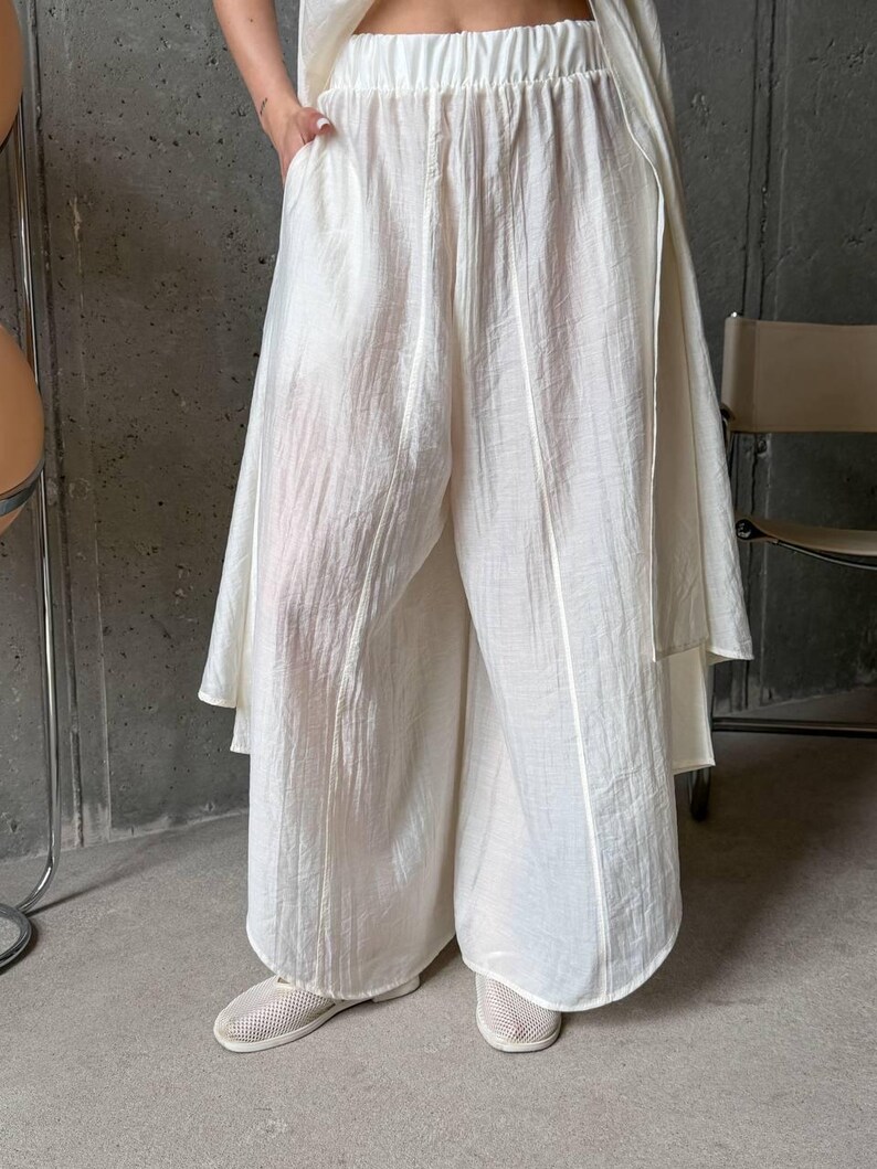 May include: Wide-leg trousers in an off-white color featuring a textured, crinkled fabric finish. These pants have an elasticized waistband, a loose, flowing silhouette, and vertical seam detailing running down the front of each leg.