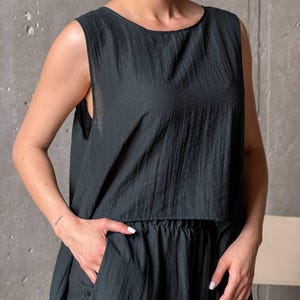 May include: A dark charcoal gray sleeveless top with a round neckline and a textured, crinkled fabric finish. The garment features a loose, boxy silhouette that falls to the waist, paired with matching dark charcoal gray bottoms with a gathered waistband.
