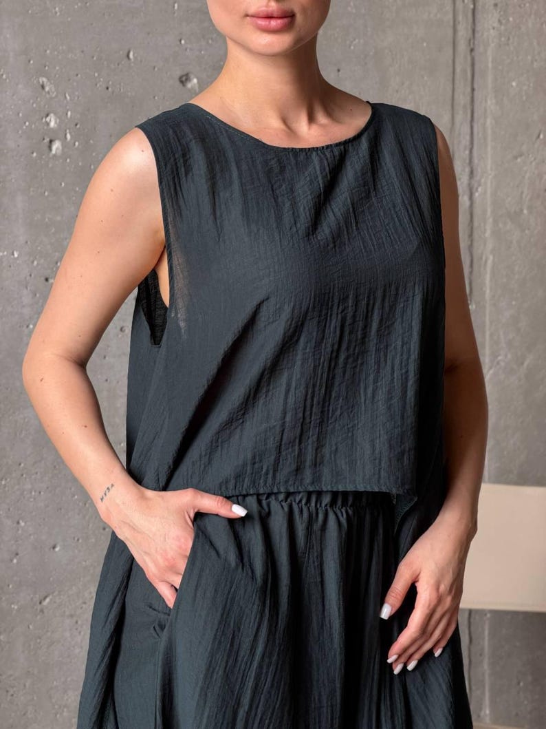 May include: A dark charcoal gray sleeveless top with a round neckline and a textured, crinkled fabric finish. The garment features a loose, boxy silhouette that falls to the waist, paired with matching dark charcoal gray bottoms with a gathered waistband.