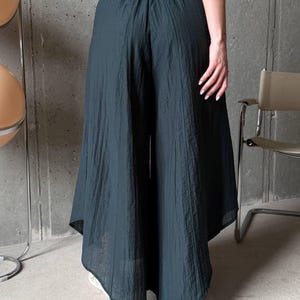 May include: Dark teal wide-leg trousers featuring a high-waisted design with an elasticated waistband. The pants are crafted from a lightweight, textured fabric that drapes loosely, creating a voluminous silhouette that extends to the lower calf.