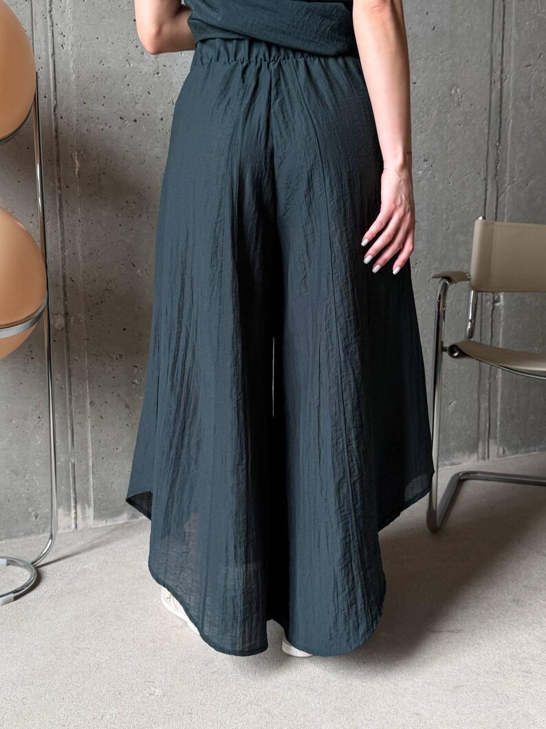 May include: Dark teal wide-leg trousers featuring a high-waisted design with an elasticated waistband. The pants are crafted from a lightweight, textured fabric that drapes loosely, creating a voluminous silhouette that extends to the lower calf.