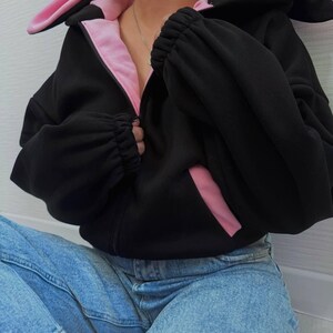 Op de afbeelding: Zwarte hoodie met ritssluiting, roze accenten en konijnenoren op de capuchon. De binnenkant van de capuchon en de zakken is roze. De mouwen zijn bij de polsen gerimpeld. Het model draagt een blauwe spijkerbroek.