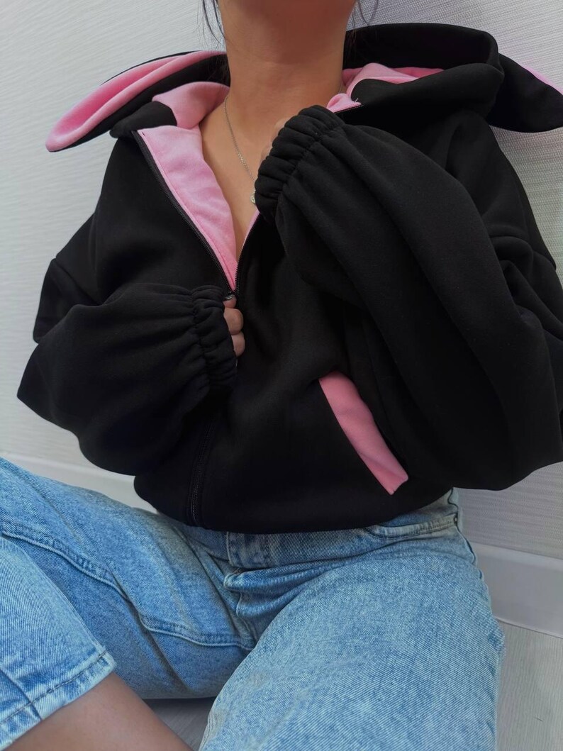 Op de afbeelding: Zwarte hoodie met ritssluiting, roze accenten en konijnenoren op de capuchon. De binnenkant van de capuchon en de zakken is roze. De mouwen zijn bij de polsen gerimpeld. Het model draagt een blauwe spijkerbroek.