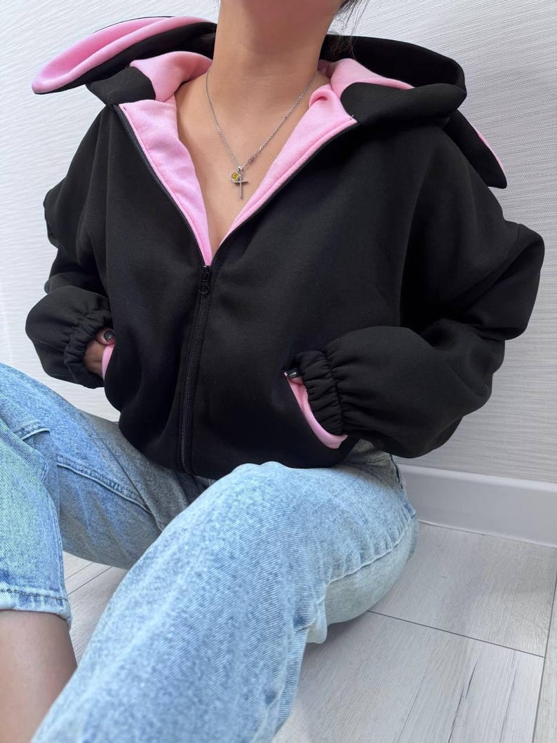 Op de afbeelding: Zwarte hoodie met ritssluiting, roze binnenkant en konijnenoordesign op de capuchon. De hoodie heeft een volledige ritssluiting, lange mouwen en roze accenten op de manchetten. De persoon draagt een lichtblauwe spijkerbroek.