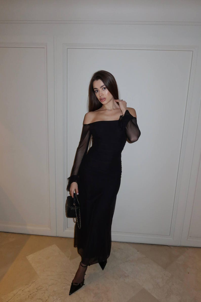 May include: A black off-the-shoulder midi dress featuring sheer long sleeves, a fitted bodice, and a straight neckline. The garment is paired with a small black handbag with a chain strap and black pointed-toe heels with ankle straps.