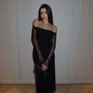 May include: A black off-the-shoulder midi dress featuring sheer long sleeves and a fitted bodice. The ensemble is paired with a small black quilted handbag with a chain strap and black pointed-toe heels, creating a cohesive monochromatic look.