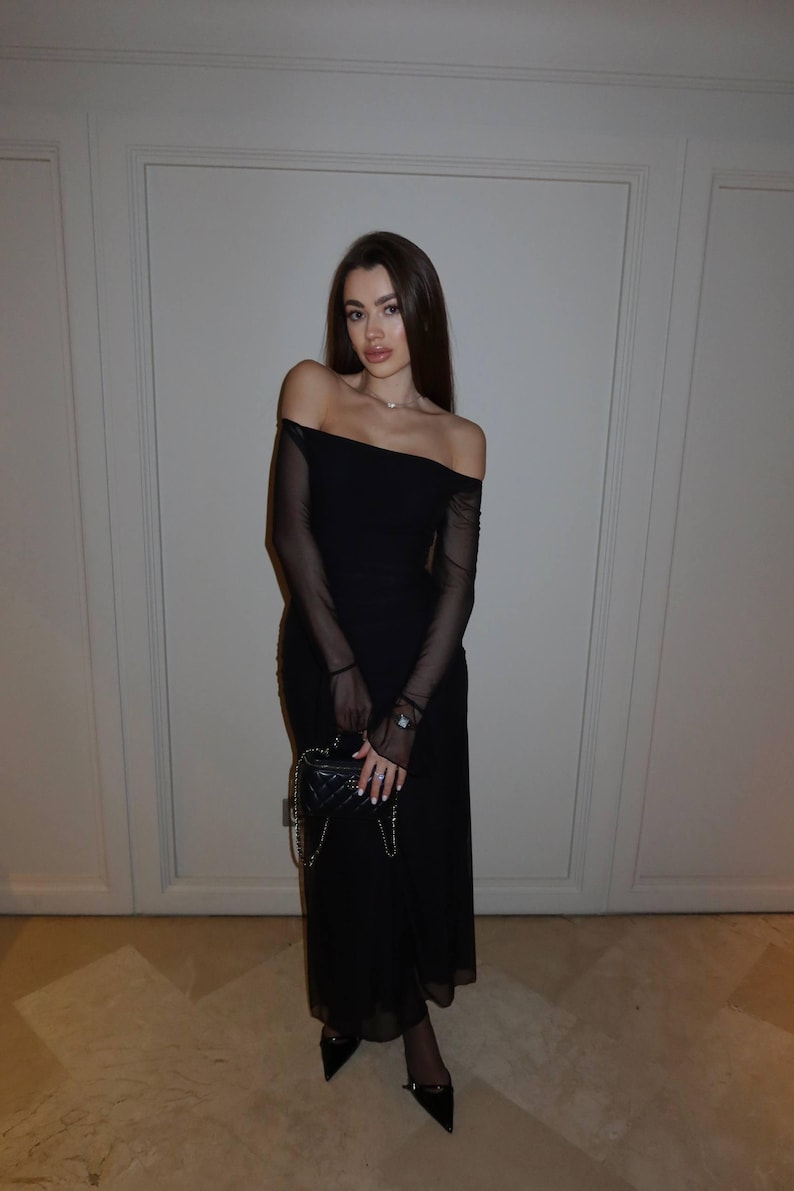 May include: A black off-the-shoulder midi dress featuring sheer long sleeves and a fitted bodice. The ensemble is paired with a small black quilted handbag with a chain strap and black pointed-toe heels, creating a cohesive monochromatic look.
