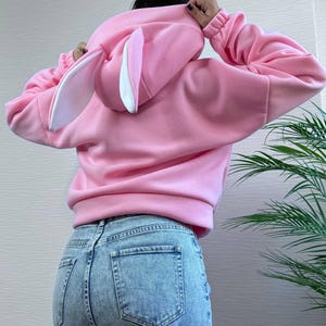 Op de afbeelding: Een lichtroze hoodie met een konijnenoordesign op de capuchon. De hoodie heeft lange mouwen en wordt gedragen met een blauwe jeans. De konijnenoren zijn wit met roze binnendetails.