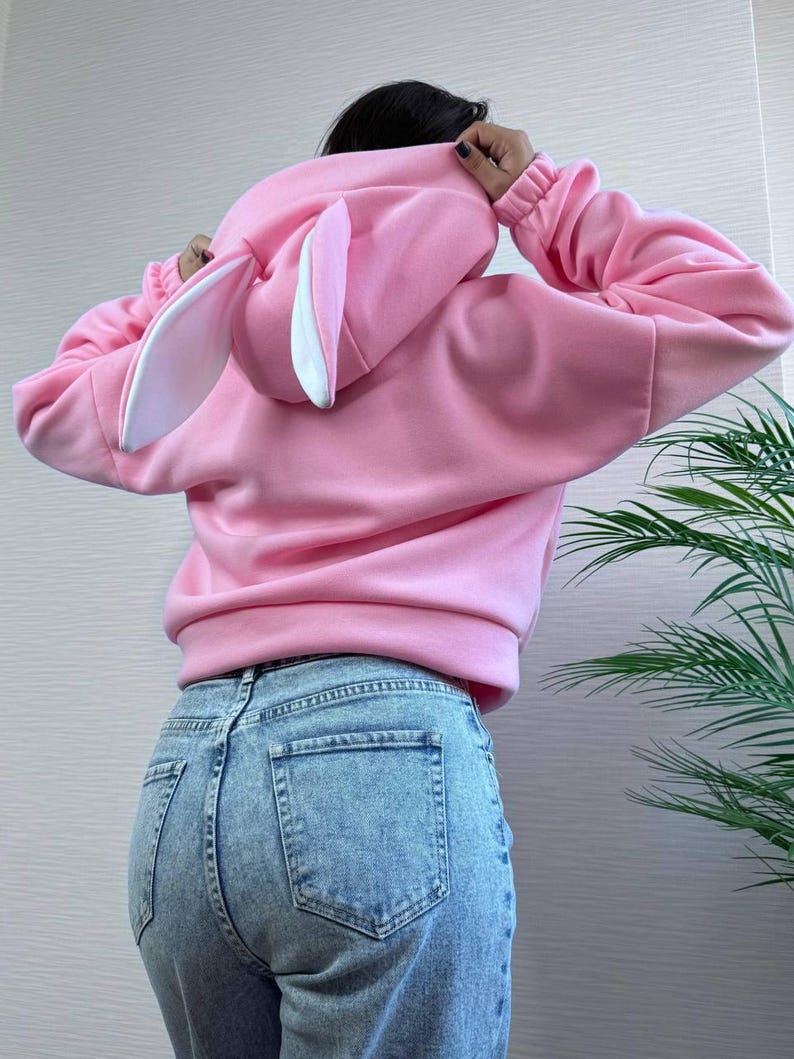 Op de afbeelding: Een lichtroze hoodie met een konijnenoordesign op de capuchon. De hoodie heeft lange mouwen en wordt gedragen met een blauwe jeans. De konijnenoren zijn wit met roze binnendetails.
