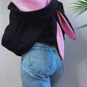 Op de afbeelding: Zwarte hoodie met roze binnenkant en lange konijnenoren. De hoodie wordt gecombineerd met lichtblauwe jeans. De oren strekken zich uit de capuchon en cre&euml;ren een speels ontwerp.
