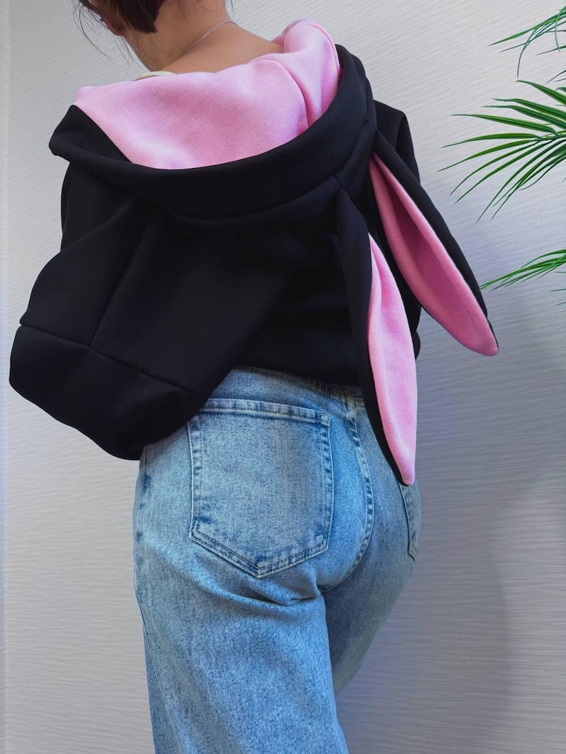 Op de afbeelding: Zwarte hoodie met roze binnenkant en lange konijnenoren. De hoodie wordt gecombineerd met lichtblauwe jeans. De oren strekken zich uit de capuchon en cre&euml;ren een speels ontwerp.