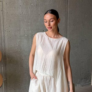 May include: A sleeveless, off-white top with a textured, crinkled fabric finish and a loose, flowing silhouette. The garment features a high, rounded neckline and a slightly cropped, asymmetrical hemline that drapes over a matching pair of trousers.