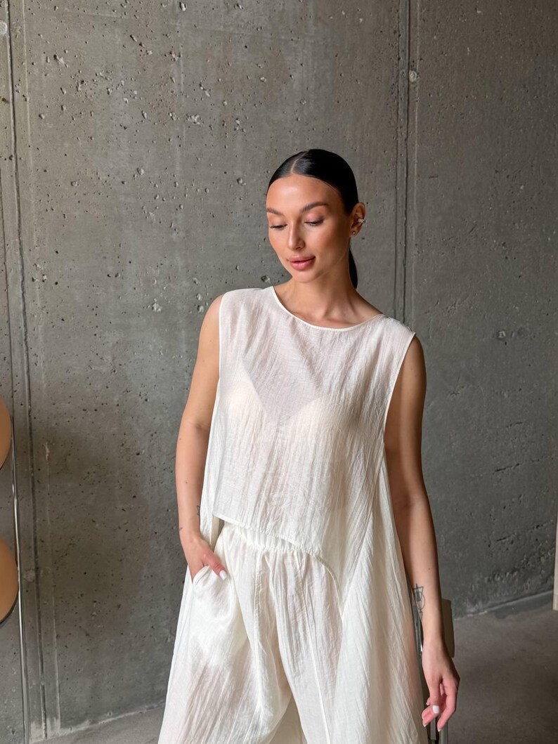May include: A sleeveless, off-white top with a textured, crinkled fabric finish and a loose, flowing silhouette. The garment features a high, rounded neckline and a slightly cropped, asymmetrical hemline that drapes over a matching pair of trousers.