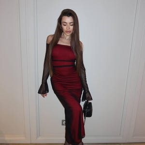 May include: A floor-length, off-the-shoulder dress in deep red with black mesh long sleeves that flare at the wrists. The dress features a fitted bodice with horizontal black detailing, a straight neckline, and a slim silhouette, paired with black pointed-toe heels and a small black handbag.
