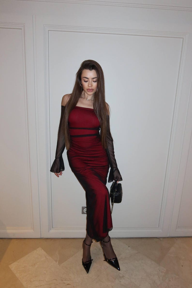 May include: A floor-length, off-the-shoulder dress in deep red with black mesh long sleeves that flare at the wrists. The dress features a fitted bodice with horizontal black detailing, a straight neckline, and a slim silhouette, paired with black pointed-toe heels and a small black handbag.