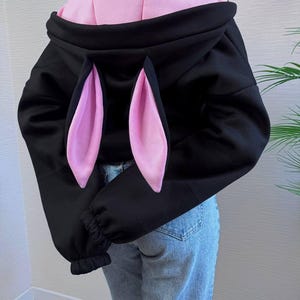 Op de afbeelding: Zwarte hoodie met roze konijnenoren en een roze binnenkant. De sweatshirt heeft lange mouwen met geribde manchetten. Het model draagt een blauwe spijkerbroek.